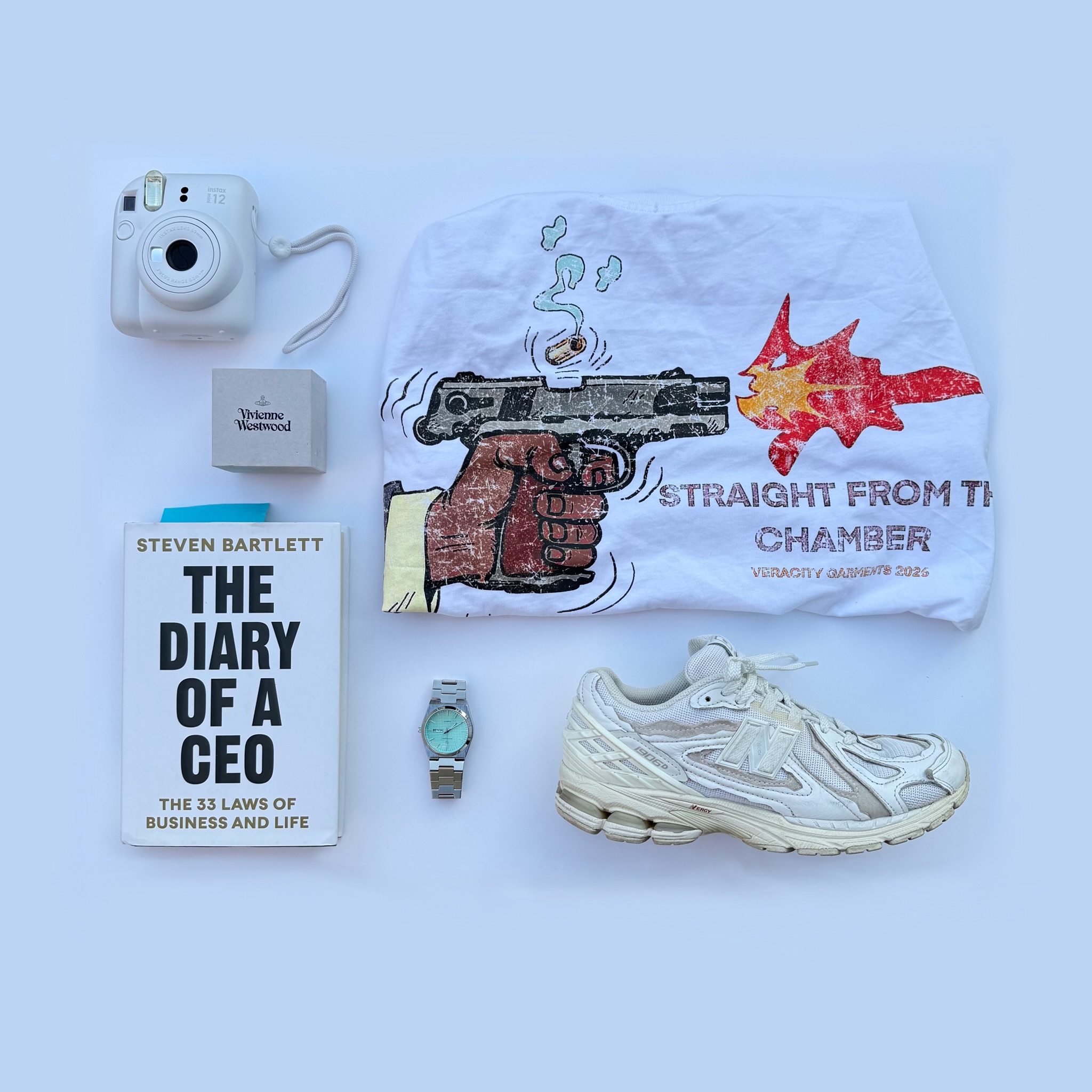 Instax camera, Steven Bartlett book, watch, and New Balance sneakers arranged on cream surface with STR8 From The Chamber Tee.